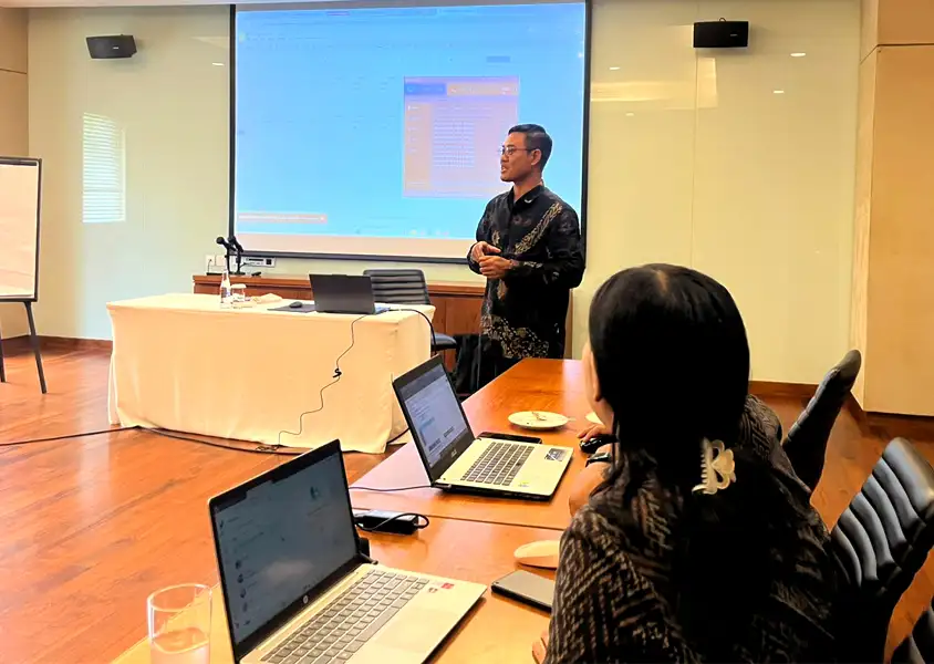 instruktur sedang berdiri menjelaskan materi Excel Intermediate yang ditampilkan melalui proyektor, sementara peserta pelatihan menyimak dengan saksama di depan